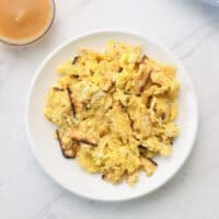 Overhead horizontal shot - a white plate with matzo brei eggs and matzo scrambled, small dishes of applesauce and sour cream beside, with a blue towel in background, on a white marble counter.