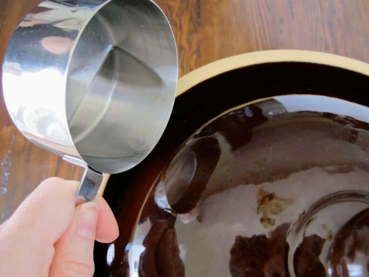 Hand with stainless measuring cup adding liquid to fermentation crock.