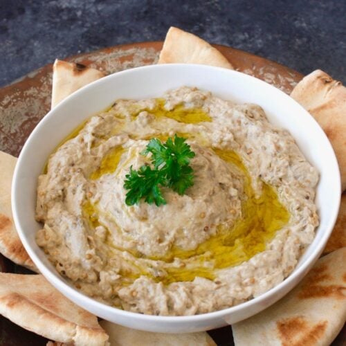A white bowl of baba ganoush drizzled with olive oil and garnished with parsley sits on a rustic platter, surrounded by slices of pita bread.
