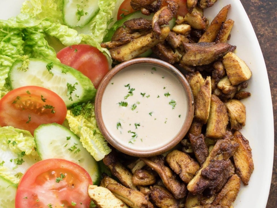 White platter of chicken shawarma with fresh lettuce and tomato, small dish of tahini in center garnished with sprinkle of parsley.