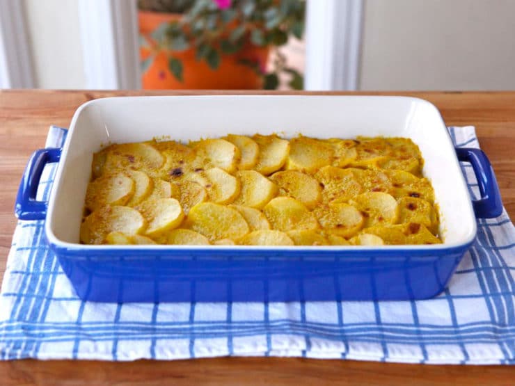 A blue casserole dish of vegan saffron scalloped potatoes on top of a white and blue striped kitchen towel.