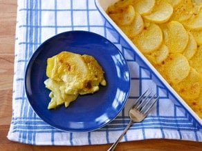 A serving of vegan saffron scalloped potatoes on a blue plate next to a casserole dish of the potatoes and a fork.