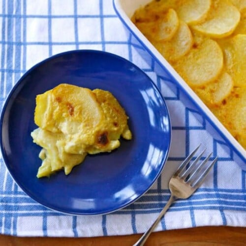 A serving of vegan saffron scalloped potatoes on a blue plate next to a casserole dish of the potatoes and a fork.