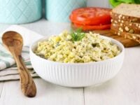 Healthy Egg Salad Pinterest Pin. A bowl of egg salad garnished with herbs, wooden spoon and cloth towel on the side, in background two aqua-colored canisters, tomato slices, bread and herbs.