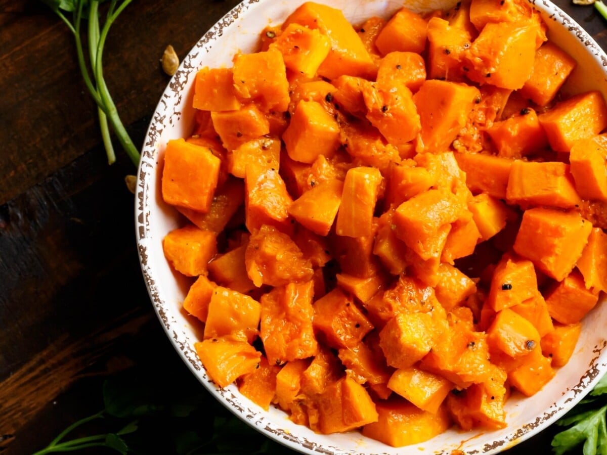 Overhead shot of a bowl filled with tender roasted butternut squash cubes on a dark wooden background, with fresh parsley and toasted butternut squash seeds scattered beside the bowl.