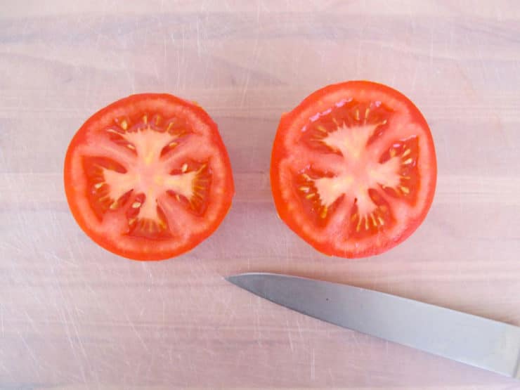 Overhead shot of tomato sliced in half with chef's knife on white plastic cutting board.