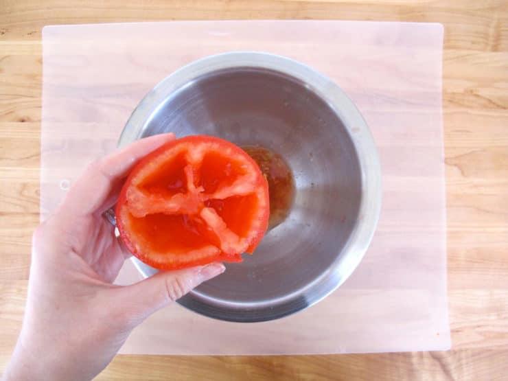 Overhead shot of stainless steel bowl, hand holding tomato half without seeds over bowl, seeds and pulp inside bowl.