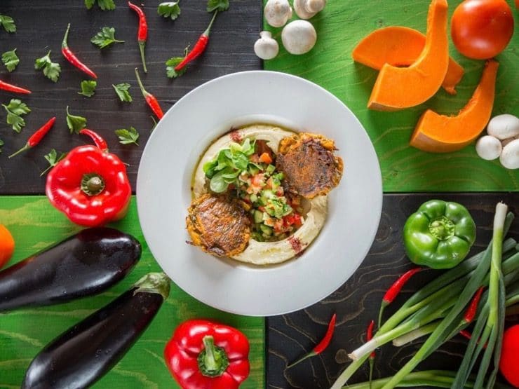 Kanaan\'s Hummus Puffer - plate of hummus topped with two baked potato pancakes and salad on green background surrounded by fresh vegetables.