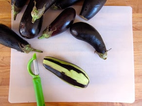 A group of whole eggplants sits on a cutting board, with one partially peeled using a green peeler, emphasizing preparation and freshness.