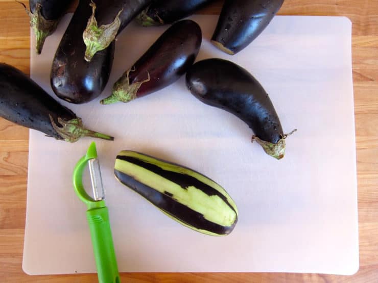 A group of whole eggplants sits on a cutting board, with one partially peeled using a green peeler, emphasizing preparation and freshness.