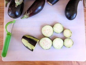 Image shows sliced eggplant on a cutting board alongside whole eggplants and a green peeler. The scene suggests preparation for cooking.