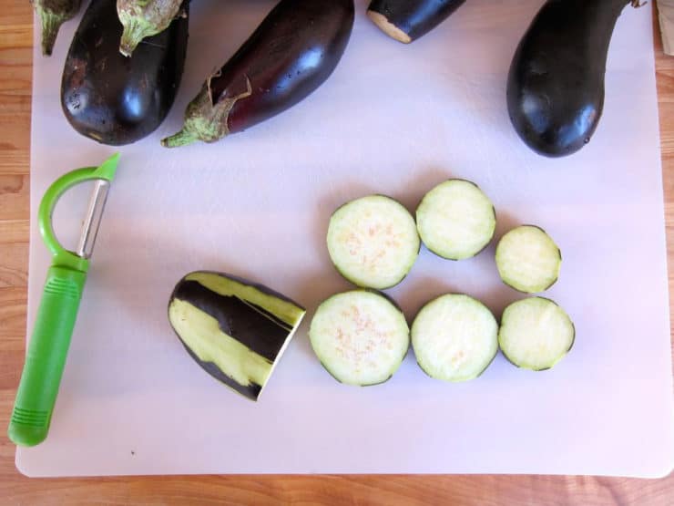 Image shows sliced eggplant on a cutting board alongside whole eggplants and a green peeler. The scene suggests preparation for cooking.