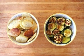 Two bowls on a wooden table: the left bowl contains seasoned, golden-brown roasted potatoes, while the right bowl holds crispy, grilled zucchini slices.