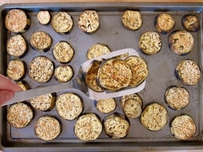 Roasted eggplant slices on a baking tray; a spatula lifts a few pieces. The slices are golden brown.