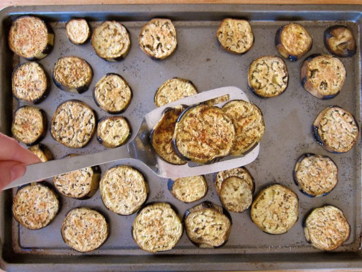 Roasted eggplant slices on a baking tray; a spatula lifts a few pieces. The slices are golden brown.