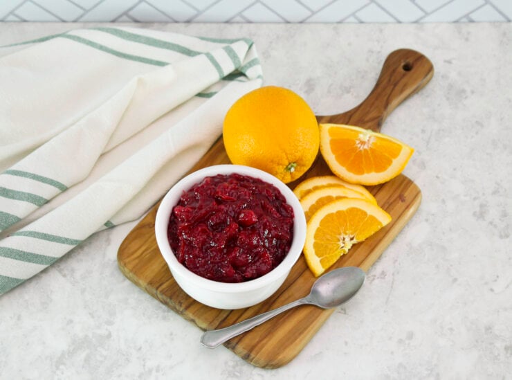 A wooden board holds a bowl of red cranberry sauce, a whole orange, orange slices and spoon. A striped towel is nearby on a light countertop.
