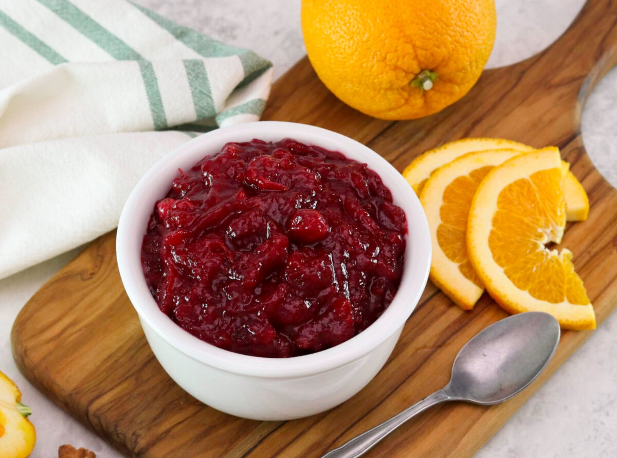 A bowl of cranberry sauce on a wooden board, surrounded by sliced and whole oranges, a spoon and a towel is draped beside it.