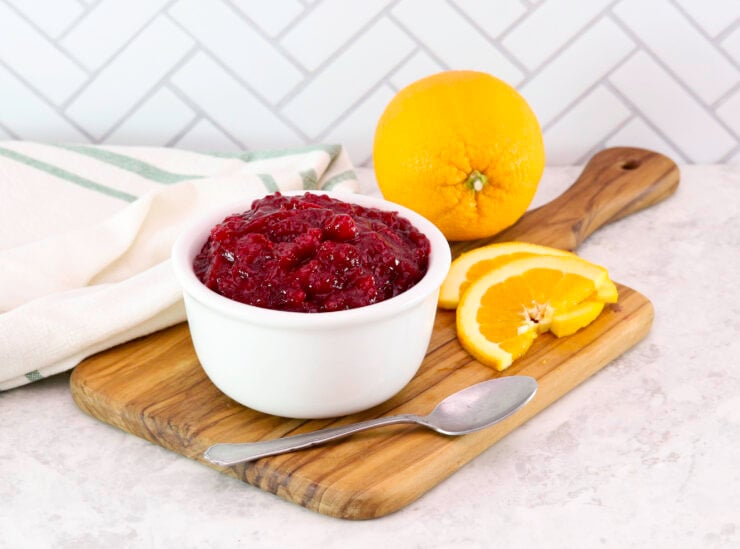 A bowl of cranberry sauce on a wooden board, surrounded by sliced and whole oranges, a spoon and a towel is draped beside it.