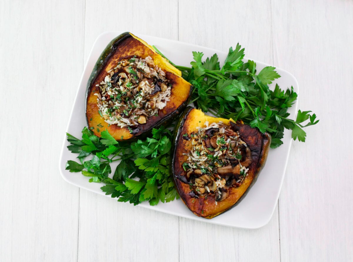 Two vegan stuffed acorn squash filled with rice, mushrooms, and herbs, garnished with fresh parsley on a white rectangular plate, on a light wooden table.Two vegan stuffed acorn squash filled with rice, mushrooms, and herbs, garnished with fresh parsley on a white rectangular plate, on a light wooden table.