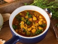 A bowl of Chickpea Kale and Fire Roasted Tomato Soup with vegetables and herbs on a cutting board