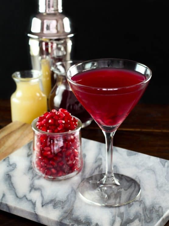 Martini glass with pomegranate martini on marble cutting board, glass carafe of pomegranate seeds, glass carafe of lime juice, martini shaker in background, on dark wooden surface.