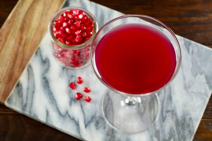Overhead view of martini glass with pomegranate martini on marble cutting board, glass carafe of pomegranate seeds, glass carafe of lime juice, martini shaker in background, on dark wooden surface.