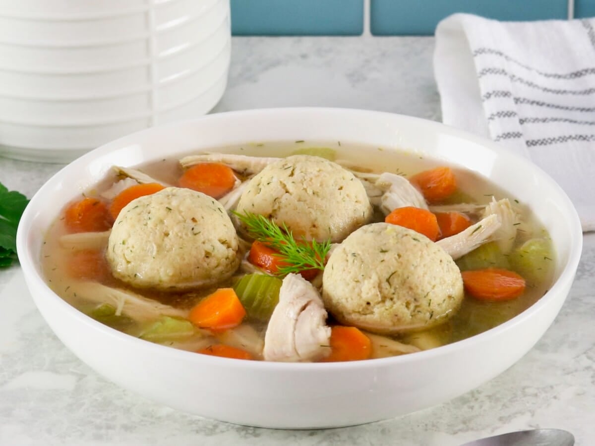 Horizontal shot of floater matzo balls in a shallow bowl of Jewish chicken soup with carrot slices, pieces of celery, and golden broth. Spoon and linen napkin on the white marble counter beside the bowl. Tiles and white jar in background.