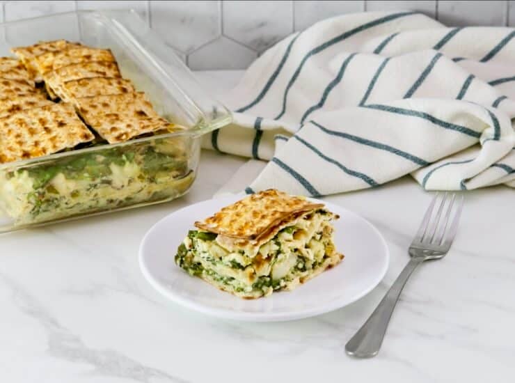 A savory spinach matzo pie in square casserole dish, a slice of spinach matzo pie with layers of spinach, feta cheese, and artichokes, baked to perfection and served on a white plate, fork and towel beside the plate. 