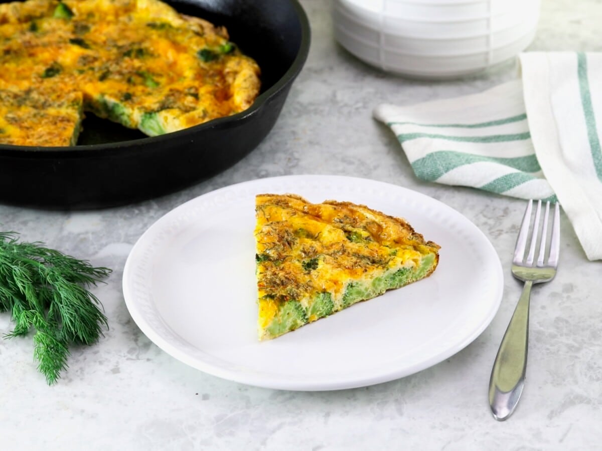 A triangular slice of broccoli cheese frittata on a small white plate in foreground with fork, larger frittata in skillet in background. Garnish of fresh dill on the side, linen napkin and canister in background.