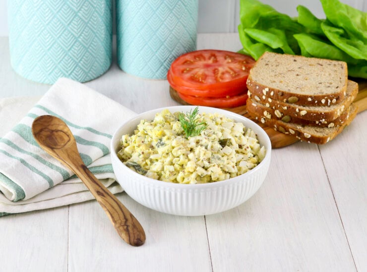 A bowl of egg salad garnished with dill sits on a wooden table. Nearby are sliced tomatoes, lettuce, seeded bread, turquoise jars, and a wooden spoon over a striped napkin.