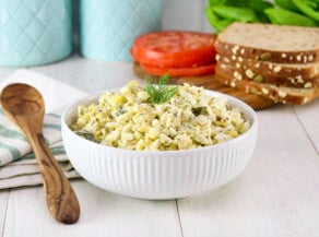 A bowl of egg salad garnished with dill sits on a wooden table. Nearby are sliced tomatoes, lettuce, seeded bread, turquoise jars, and a wooden spoon over a striped napkin.
