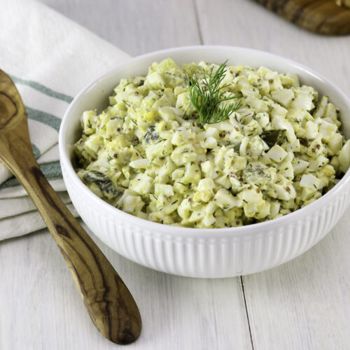A bowl of egg salad garnished with dill sits on a wooden table. Nearby are striped napkin, and a wooden spoon.