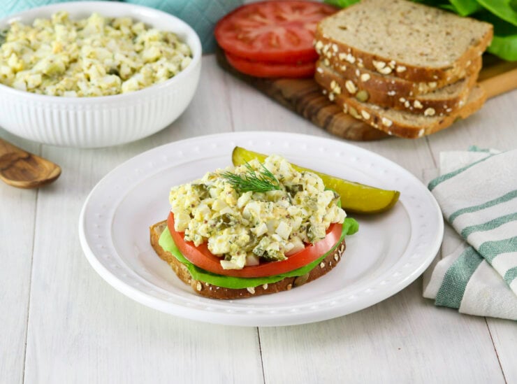 Open-faced egg salad sandwich with tomato and lettuce on whole grain bread, garnished with dill and served with a pickle slice on a white plate. Nearby are a bowl of egg salad, striped napkin, sliced tomatoes, lettuce, seeded bread and a wooden spoon.