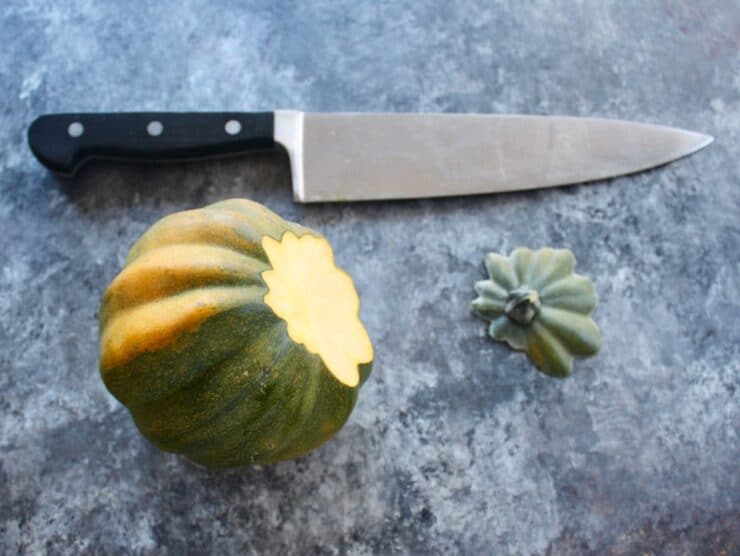 Acorn squash, stem end sliced off, stem end beside it with chef's knife above on concrete surface.