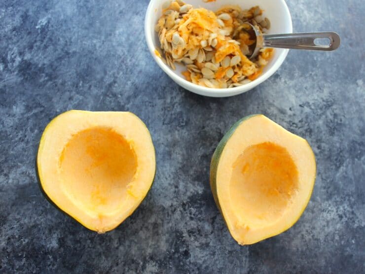 Two acorn squash halves with bowl of scooped seeds and pulp, tablespoon in background.