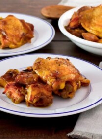 White plate of Israeli Chicken Sofrito with potatoes, fork and napkin on the side, large dish of chicken and another smaller dish in the background, on a wooden table.