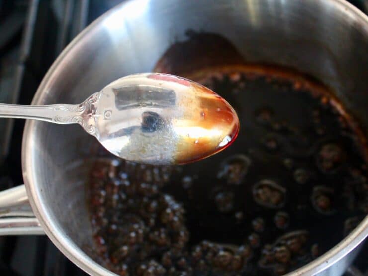 Spoon upside down showing thickened balsamic coating the back of the spoon, small saucepan with bubbling balsamic reduction sauce on stovetop in background.