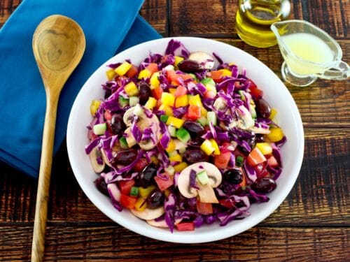 Overhead shot of bowl of colorful rainbow salad with peppers, cucumbers, mushrooms and cabbage in a white bowl with glass dish of lemon juice, carafe of olive oil and blue napkin with wooden spoon in the background.