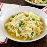 A bowl of chicken with homemade noodles and sweet peas with cloth napkin and spoon on wooden background.
