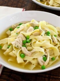 A bowl of chicken with homemade noodles and sweet peas with cloth napkin and spoon on wooden background.