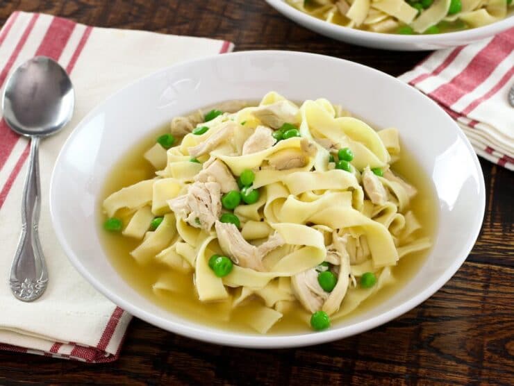 A bowl of chicken with homemade noodles and sweet peas with cloth napkin and spoon on wooden background.