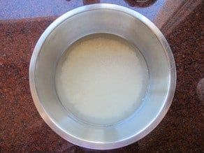 A metal bowl containing uncooked rice soaking in water, placed on a brown speckled countertop.