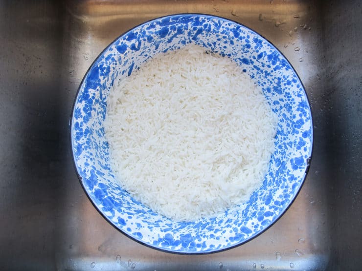 A blue speckled bowl filled with white rice sits in a stainless steel sink. The rice appears rinsed, with a clean and fresh appearance.