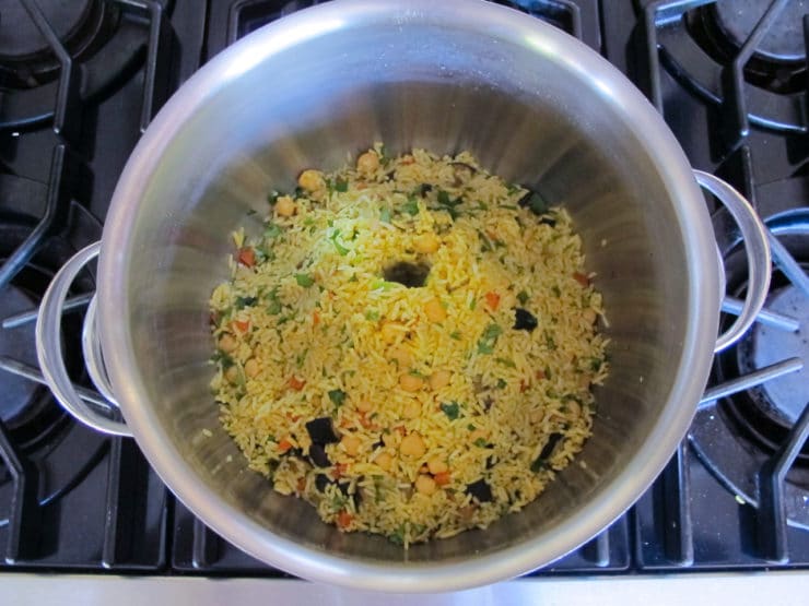 A stainless steel pot on a stove filled with colorful rice pilaf, containing visible chickpeas, carrots, and herbs.