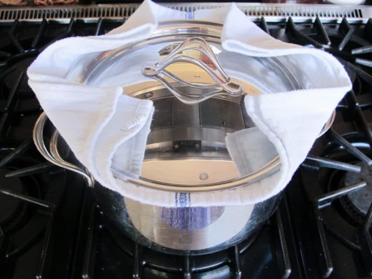 A stainless steel pot with a lid sits on a stove, covered by a white towel.