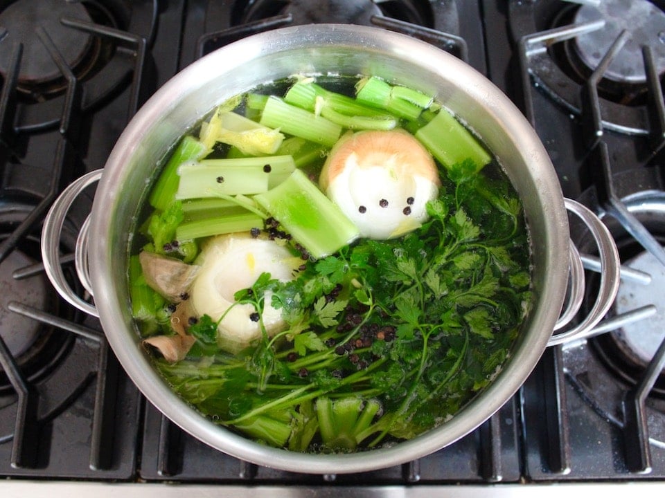 Pot of chicken stock cooking with onion, parsley, celery and peppercorns in large pot on stovetop.