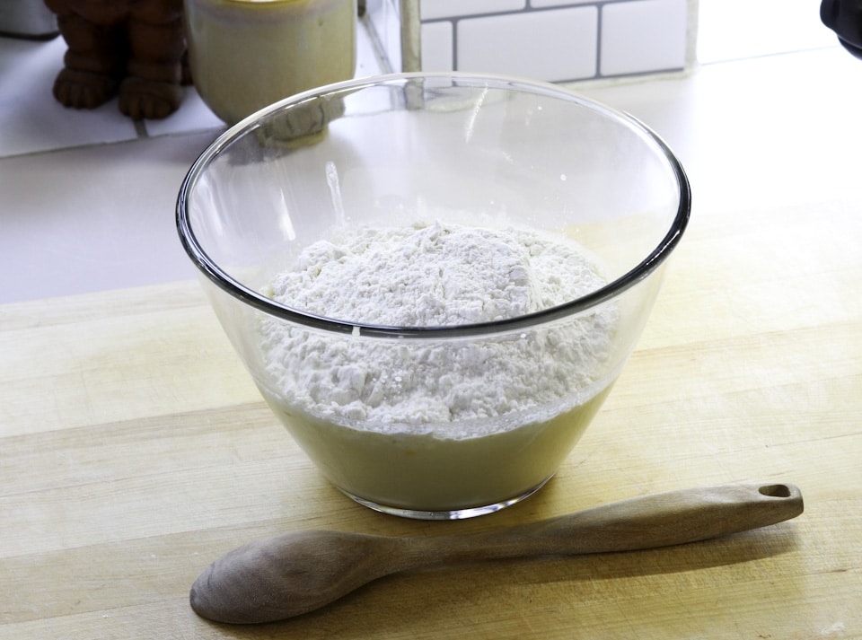A glass bowl filled with flour rests on a wooden surface, beside a wooden spoon.