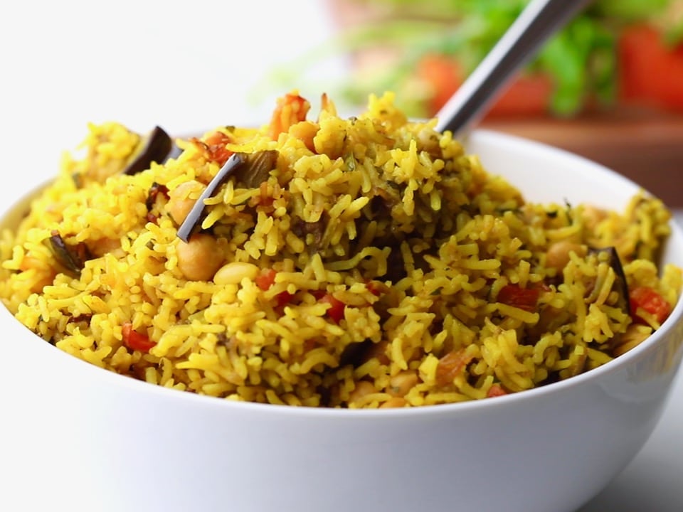 A spoon digging into fluffy yellow vegetable rice with pieces of eggplant and carrot.