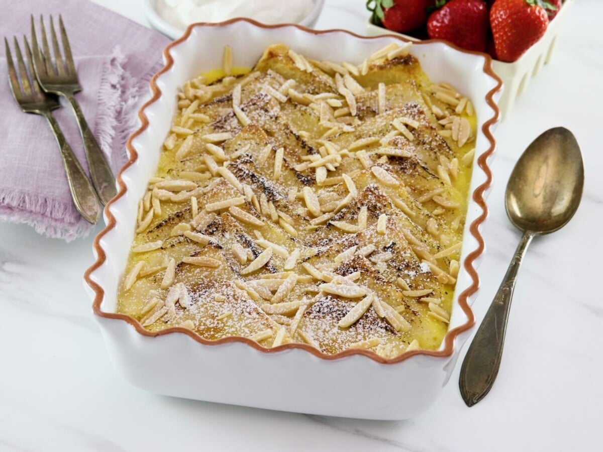 A scalloped baking dish filled with Princess Diana's favorite bread and butter pudding recipe, with spoon, forks, and linen napkins. Strawberries in background.