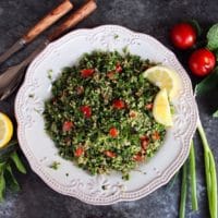 Overhead shot of quinoa tabbouleh salad in a white dish with lemon slices, half lemon, mint sprigs, green onions and small tomatoes. Salad fork and spoon on the upper left of the dish.